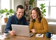 Couple in their 30s sitting at a wooden table studying a laptop together, smiling and laughing. A mug on the left has the words "ADVENTURE AWAITS". This photo represents students or aspiring mortgage professionals utilizing NMLS test prep materials like practice questions and study guides, preparing to pass the NMLS SAFE MLO exam and start a new career.